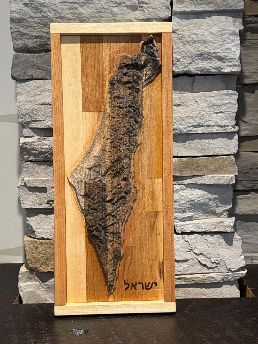 A rectangular 3D carved wood map of Israel, handcrafted from light maple and warm oak hardwoods. The detailed topographical map showcases the distinct geographical regions and textures of the land, mounted vertically on a smooth wooden backboard with a clean border. __etsy:8156430338